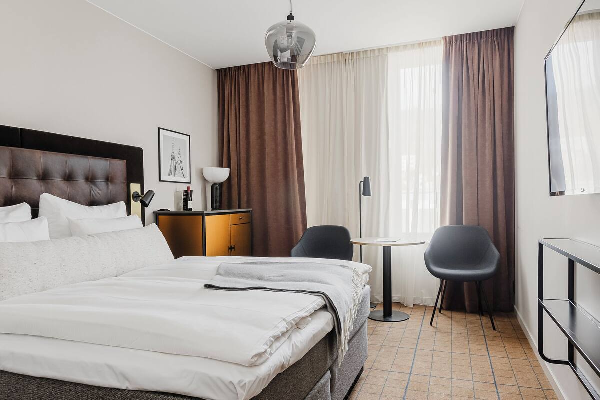 A hotel room with a large bed, a desk, two chairs, a lamp, and a picture on the wall.