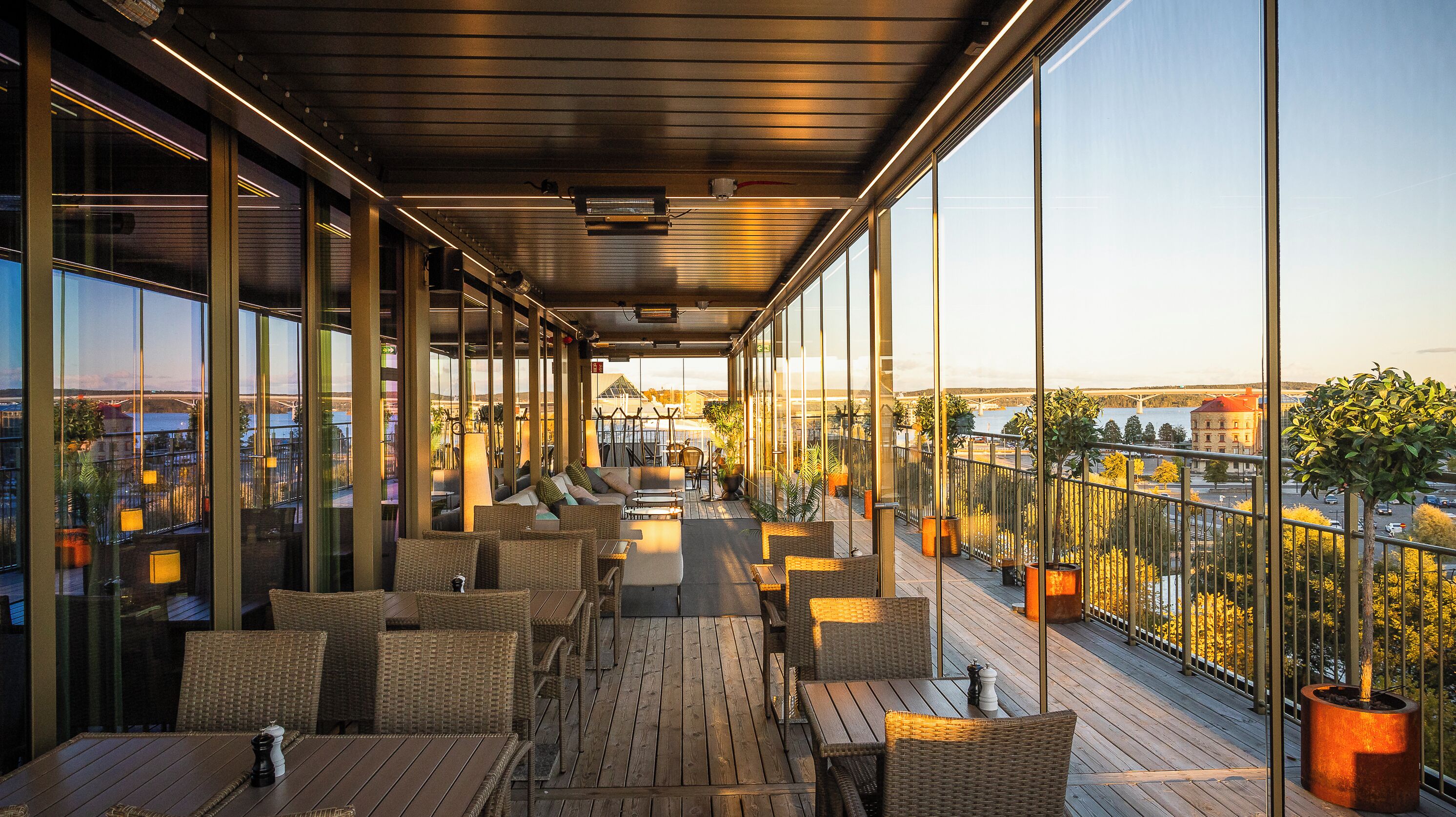 Een ruim balkon met rieten meubilair, grote ramen met uitzicht over de stad, en plantenpotten.