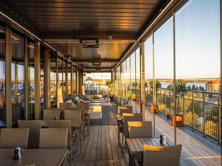 A spacious balcony with wicker furniture, large windows offering a city view, and potted plants.