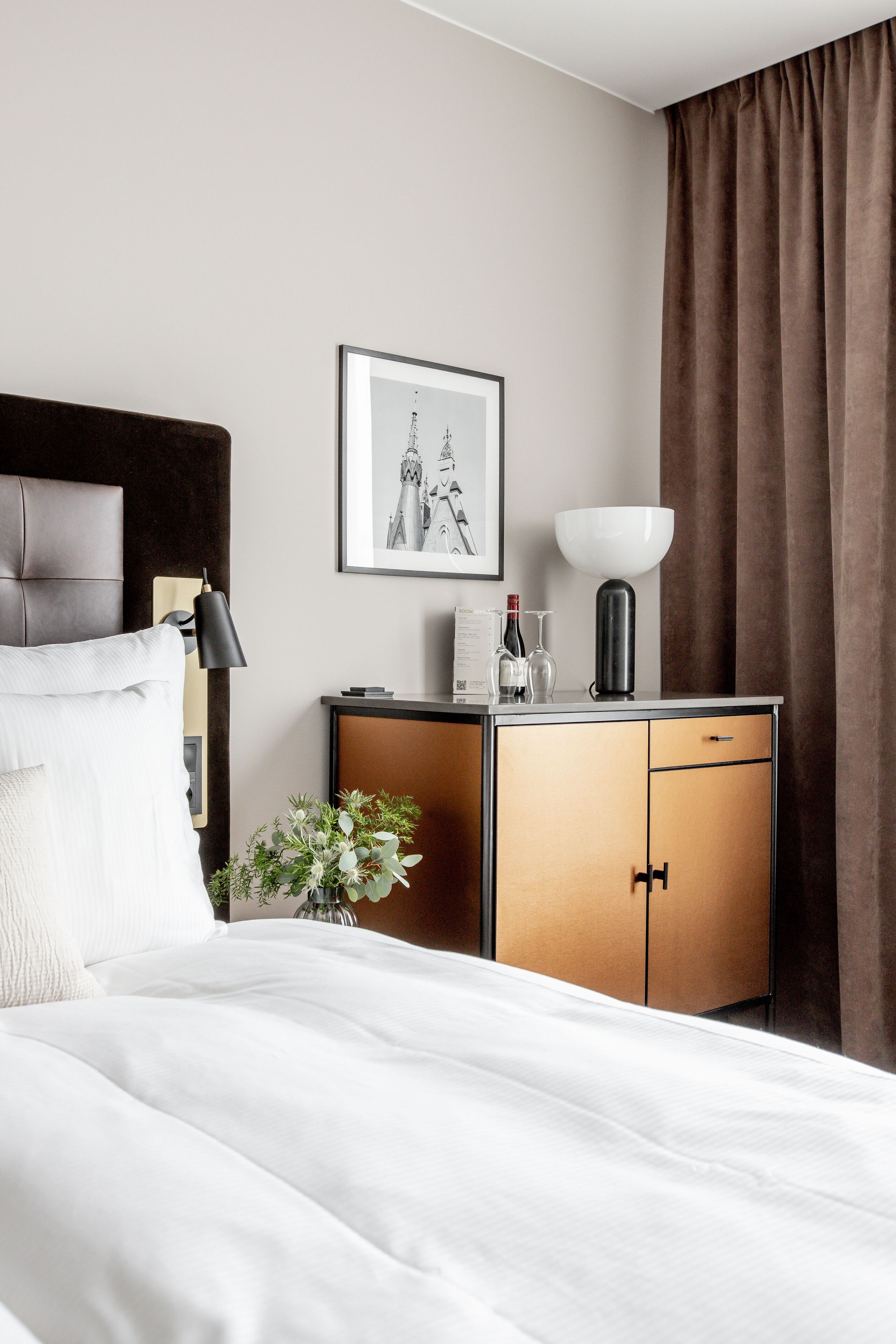 A hotel room with a bed, a bedside table with a lamp, a framed picture on the wall, a cabinet with a bowl, and a window with curtains.