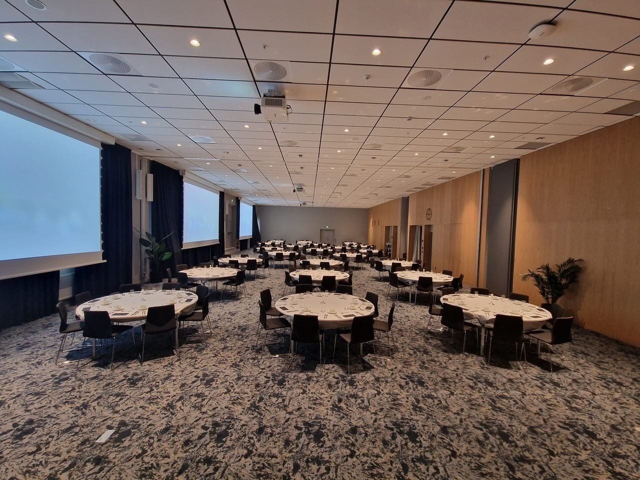A conference room with multiple round tables, carpeted flooring, and a projector screen on the wall.