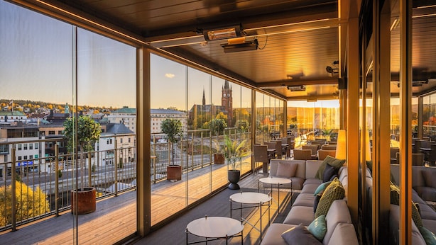 A modern outdoor seating area with large windows, a view of a cityscape, and a wooden deck. - Clarion Hotel Sundsvall (Sundsvall)