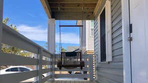 Une terrasse en bois avec une chaise suspendue, une porte blanche et une vue sur une voiture et des arbres.