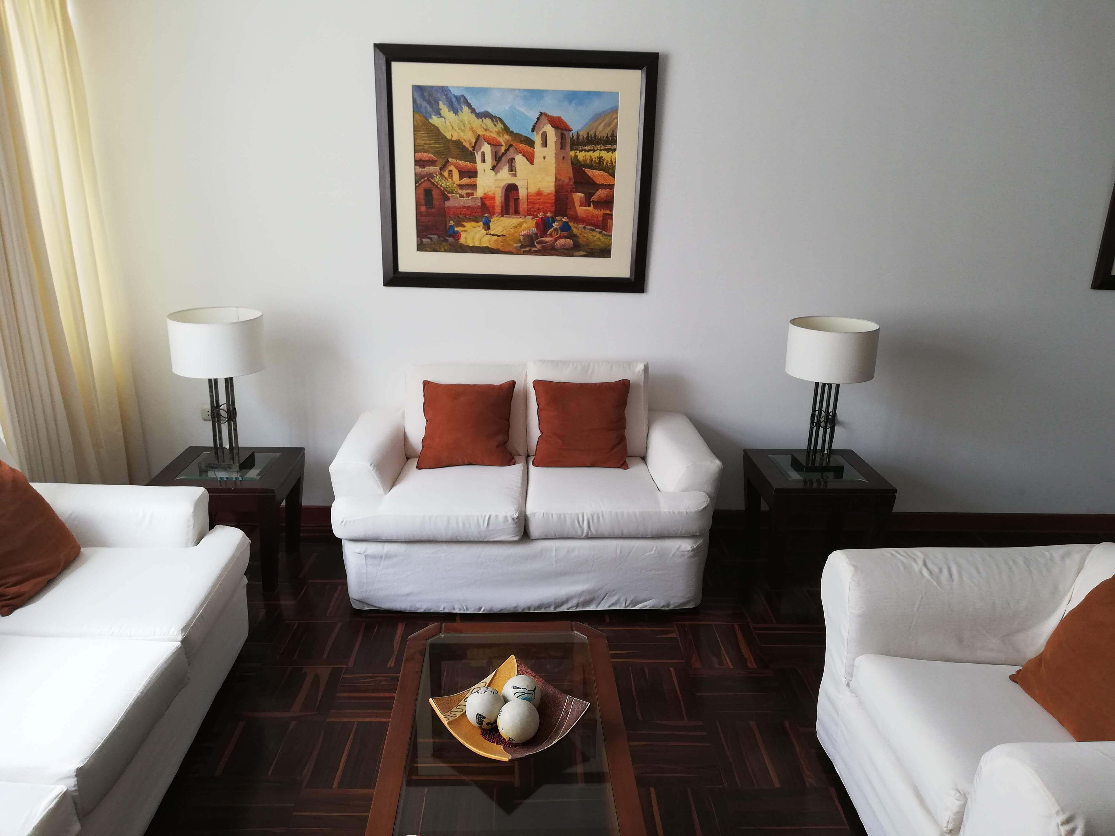 A hotel room with a white sofa set, two side tables with lamps, a coffee table with decorative items, and a framed painting on the wall.