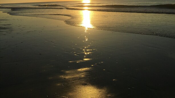 Ein Strandbild mit untergehender Sonne über dem Meer, deren Licht sich im feuchten Sand spiegelt.