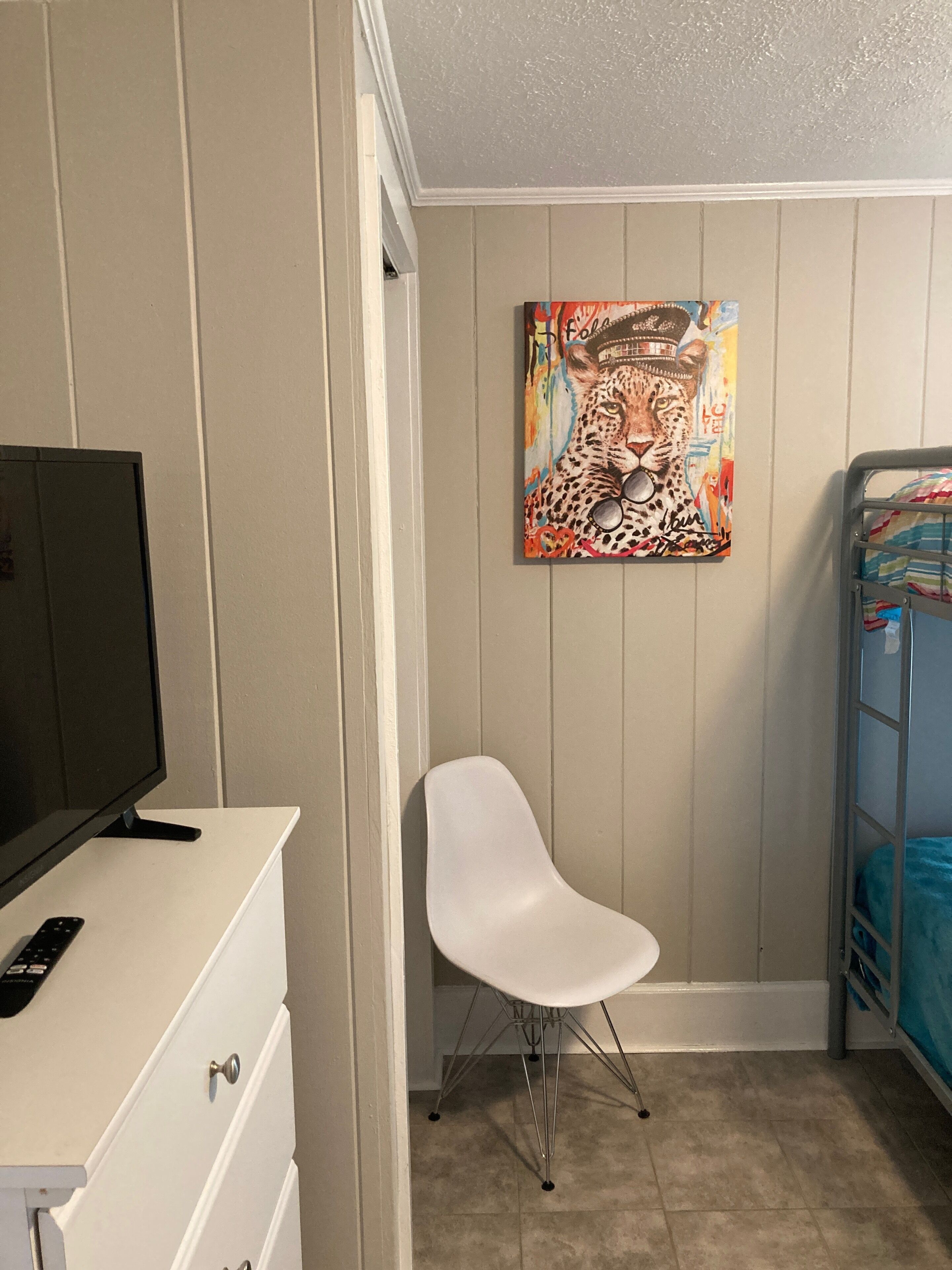 A bedroom with a TV, a white chair, a bunk bed, and a colorful leopard painting on the wall.
