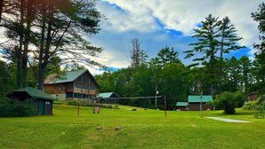 A rustic cabin with a green roof, surrounded by a grassy field and dense trees. - Chalet Cabin,  Gore Skiing, Pool, AC, Spacious Cabin, Loon Lake (Chestertown)