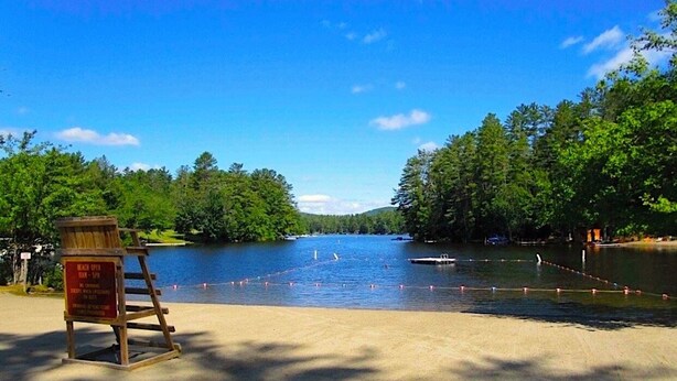 A beach with a wooden sign, a sandy shore, and a calm lake surrounded by trees. - Chalet Cabin,  Gore Skiing, Pool, AC, Spacious Cabin, Loon Lake (Chestertown)