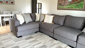 Un salon moderne avec un canapé d’angle gris, un tapis à motifs et un tableau représentant un paysage, encadré.
