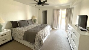 Une chambre à coucher comprenant un lit, un ventilateur de plafond, une télévision, une commode et une fenêtre avec des rideaux.