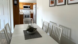 Un coin repas avec une table blanche, des chaises et une plante en guise de décoration centrale. On aperçoit une cuisine à l’arrière-plan, avec des meubles et des appareils en bois.