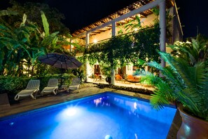 Ein Swimmingpool mit Liegestühlen und Sonnenschirm, umgeben von üppiger Vegetation und einem Gebäude mit einem Ziegeldach.