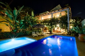Piscina con sillas de descanso y una sombrilla, rodeada de exuberante vegetación e iluminación exterior en un edificio.