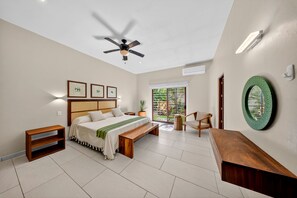Un dormitorio con cama, ventilador de techo, banca, silla, mesita de noche y ventana con vista a zonas verdes.