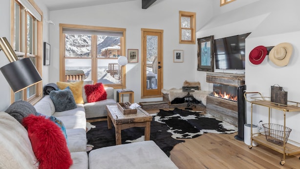 Un salon chaleureux avec une cheminée, un canapé agrémenté de coussins colorés, un tapis en peau de vache et une vue sur des montagnes enneigées par de grandes fenêtres.