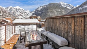 Une terrasse en bois avec un foyer, des bancs et une vue sur des montagnes enneigées.