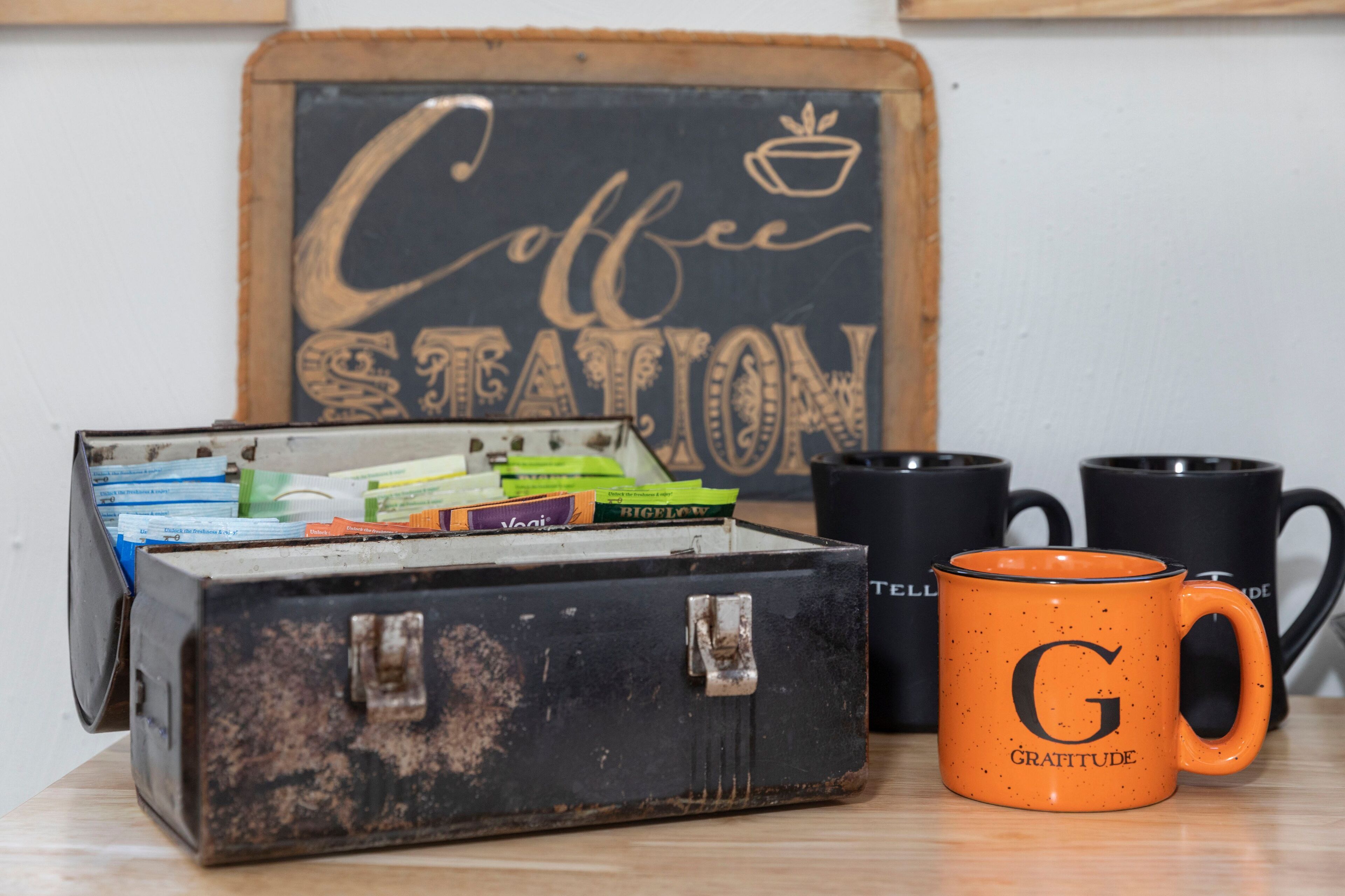 A rustic metal box filled with coffee packets, a black mug, and an orange mug with a letter on it, all on a wooden surface.