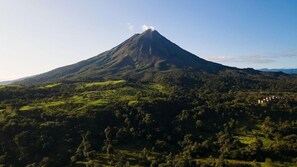 A mountain with a peak emitting smoke, surrounded by lush green hills and a small settlement in the valley. - Campo Verde (La Fortuna)