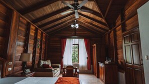 A log cabin interior with a high ceiling, wooden walls, a bed, a desk, a chair, a window with curtains, and a ceiling fan. - Campo Verde (La Fortuna)