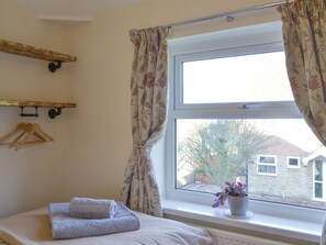 A bedroom with a bed, a window with curtains, a radiator, a plant, and wooden hangers on a wall.