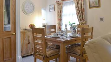 A dining area with a wooden table set for four, chairs with cushions, a clock on the wall, and a window with curtains.
