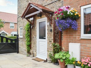 Une petite maison en briques avec une porte blanche, un porche en bois et des jardinières suspendues.