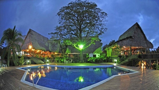 Zona de piscina con terraza de madera, un árbol grande y edificios con techo de paja.