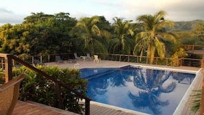 Un área de piscina con terraza de madera, sillas de descanso y palmeras.