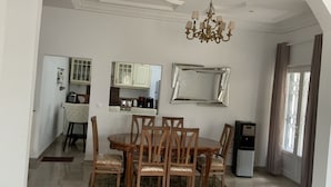 A dining area with a wooden table and chairs, a chandelier, a water dispenser, and a kitchen in the background.
