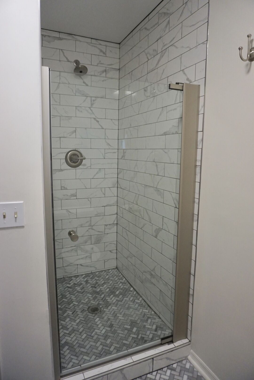 A corner shower with white marble tiles and a herringbone-patterned floor.