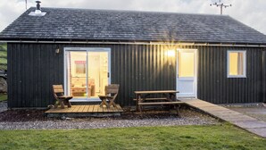 A small, dark-colored building with a sloped roof, a wooden deck with two chairs and a table, and a door leading to an interior space.