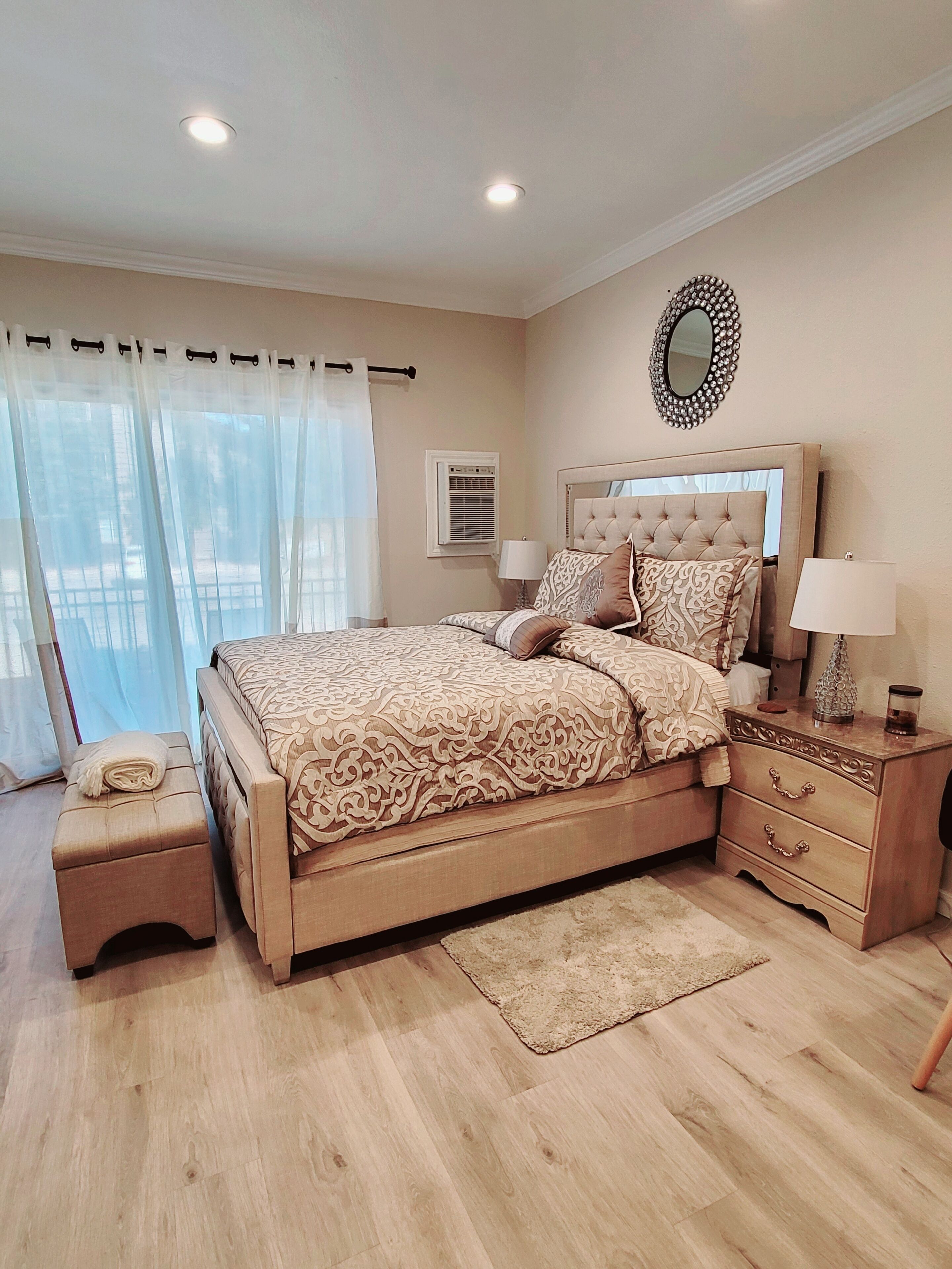 A bedroom with a large bed, a bedside table, a nightstand, a mirror, and a window with curtains.