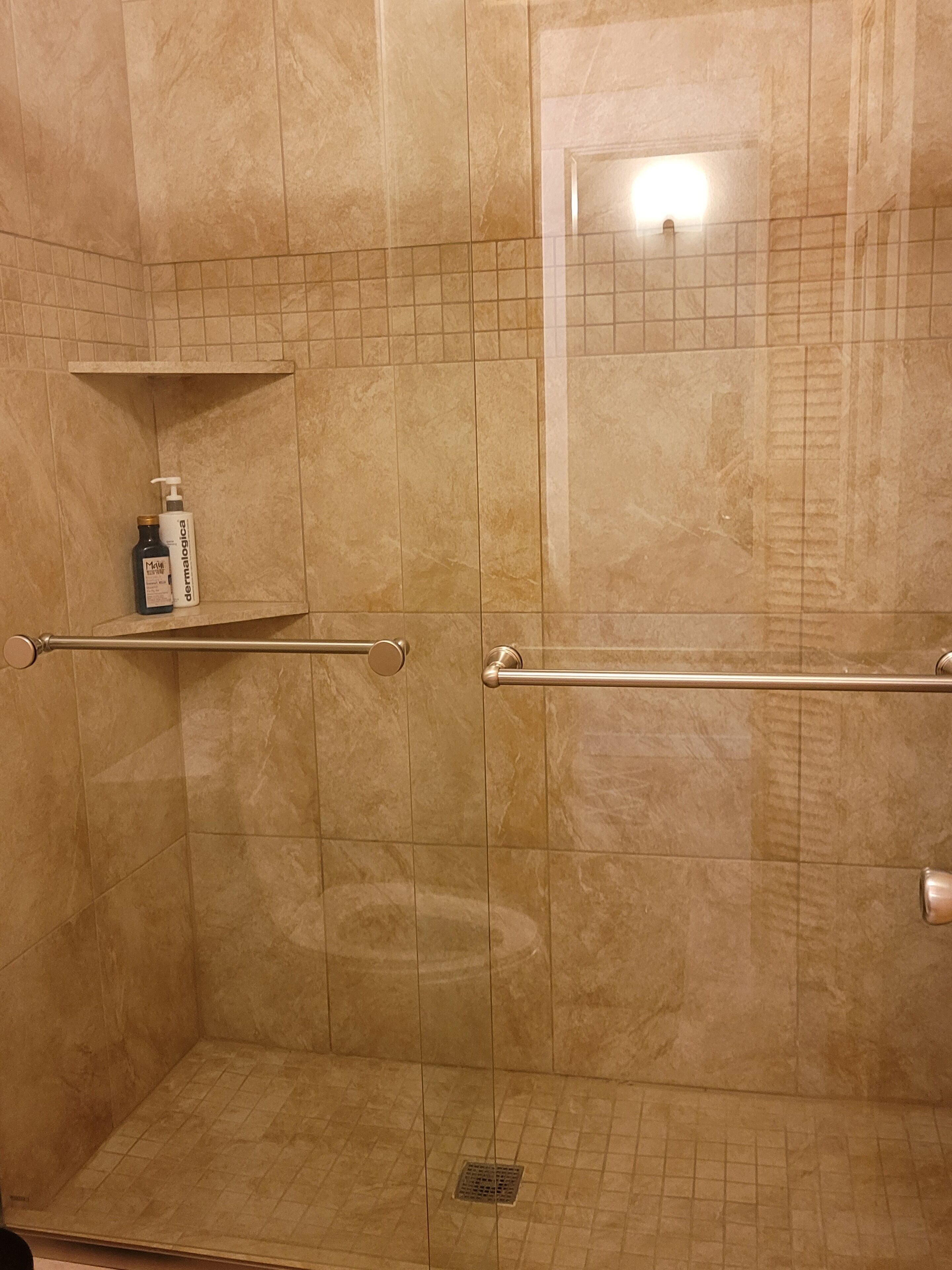 Et bruseområde med beige fliser, et gelænder, en hylde med toiletartikler og et lille spejl.