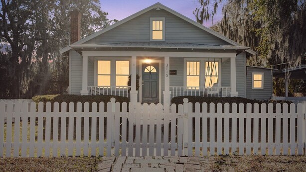 Een tweelaags huis met een witte picket fence, een veranda en een oprit van baksteen.