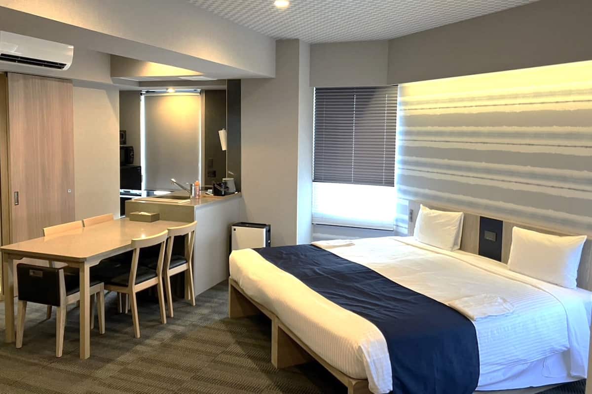 Hotellihuone, jossa on sänky, työpöytä, tuoli, peili ja ikkuna, jossa on sälekaihtimet.