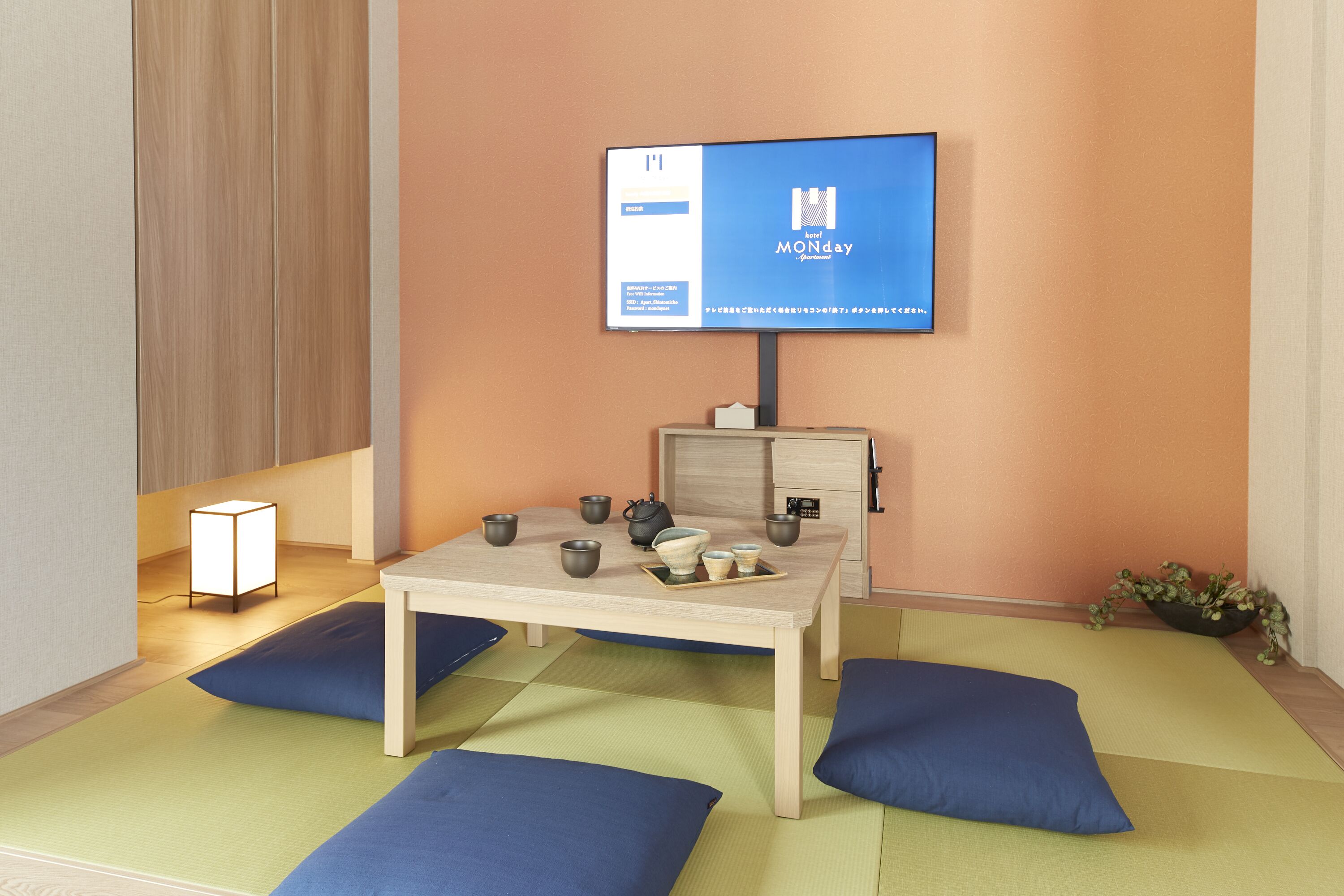 Een hotelkamer met een houten tafel, blauwe kussens, een televisie waarop de naam van het hotel staat en een kleine plant aan de zijkant.