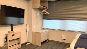 A hotel room with a flat-screen TV, a desk with a chair, a window with blinds, a vacuum cleaner, and a wardrobe.