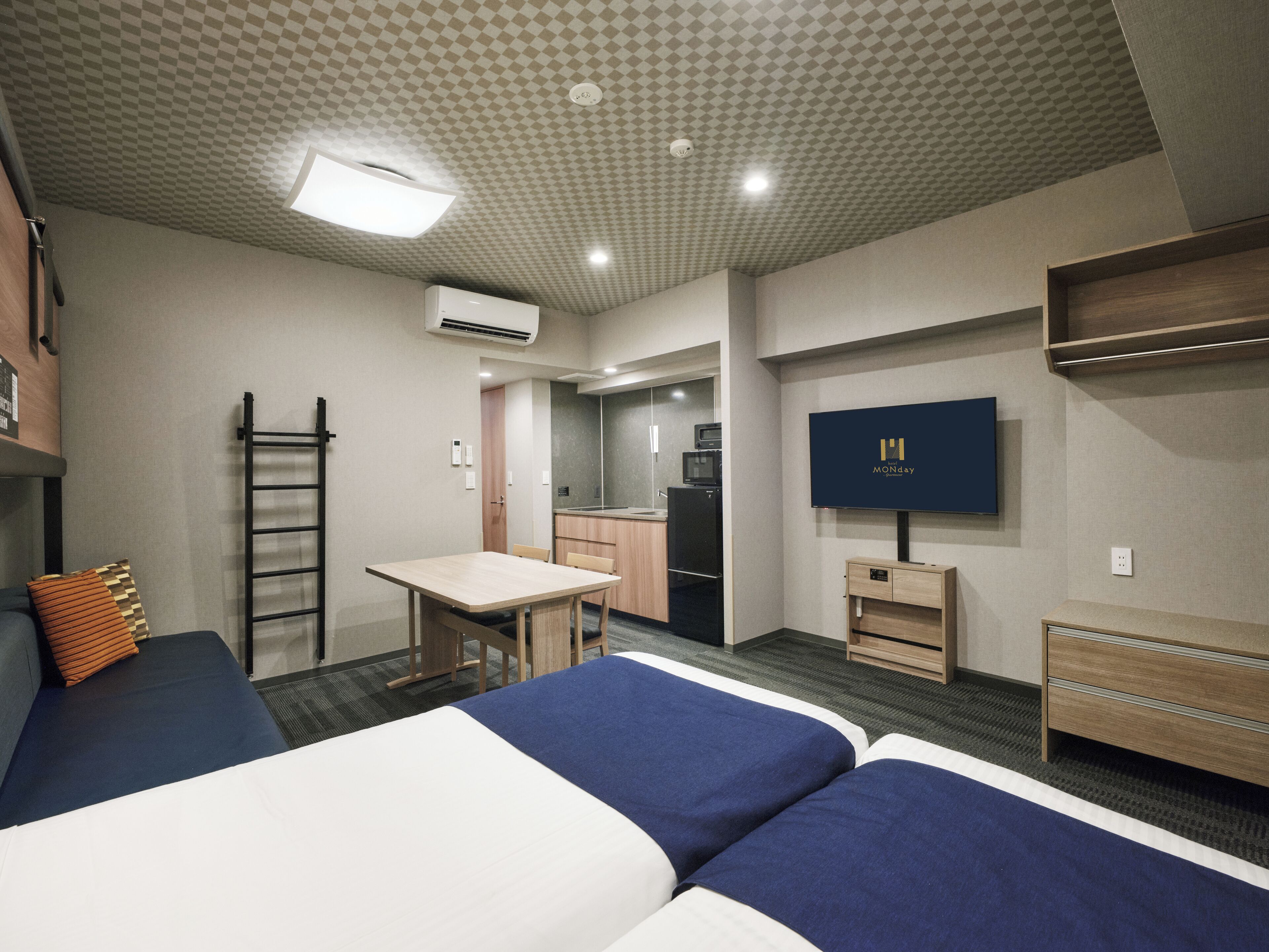 A hotel room with two beds, a desk, a kitchenette, and a TV.