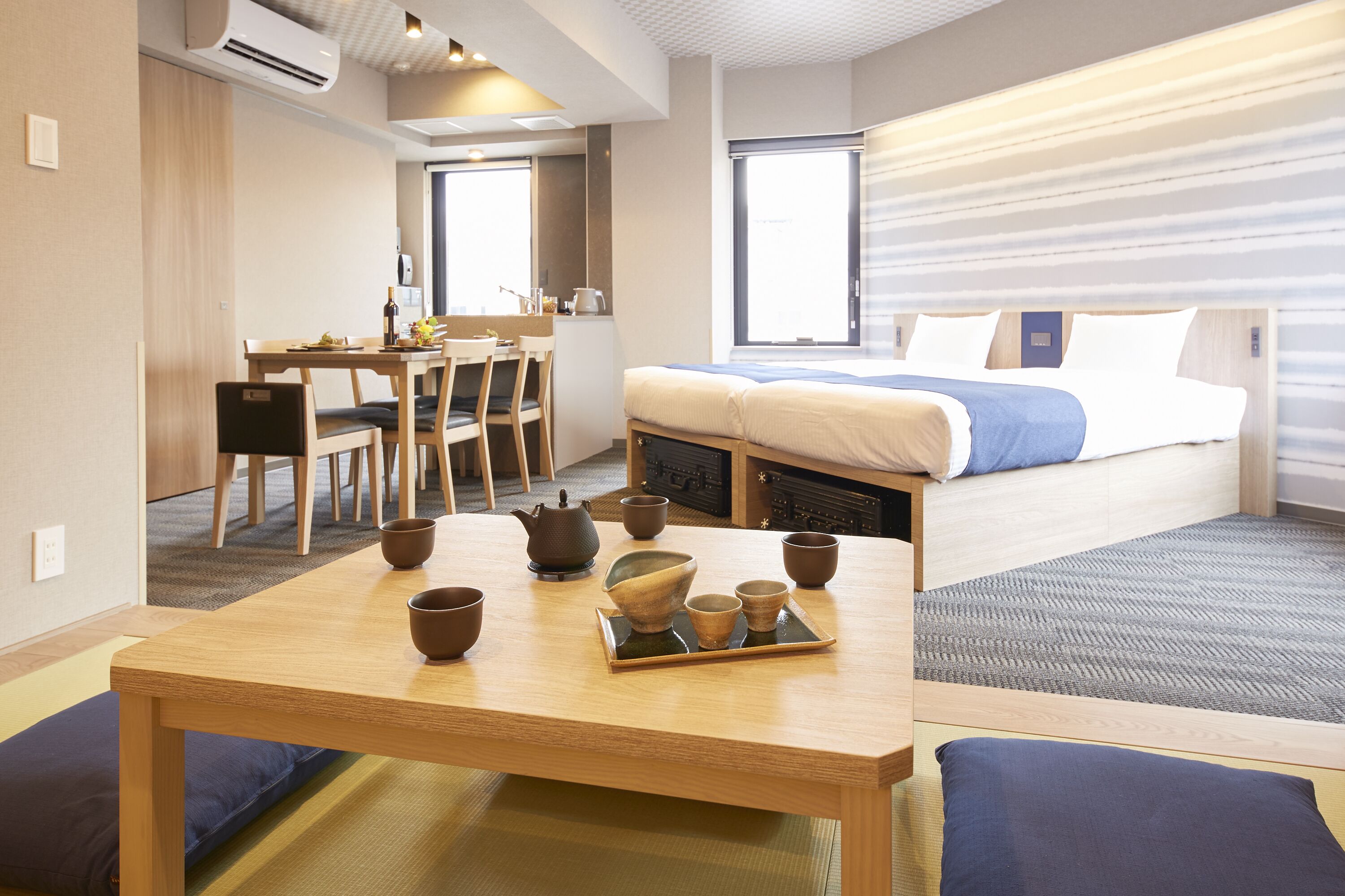 A hotel room with a bed, a dining table with chairs, a kitchen area, and a view of the outside.