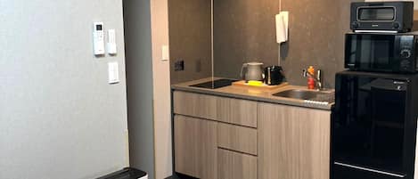 Una cocina compacta equipada con microondas, tostadora y refrigerador. Incluye un lavamanos pequeño y una encimera con hervidor. El espacio presenta un diseño moderno, con gabinetes de madera clara y pared gris.