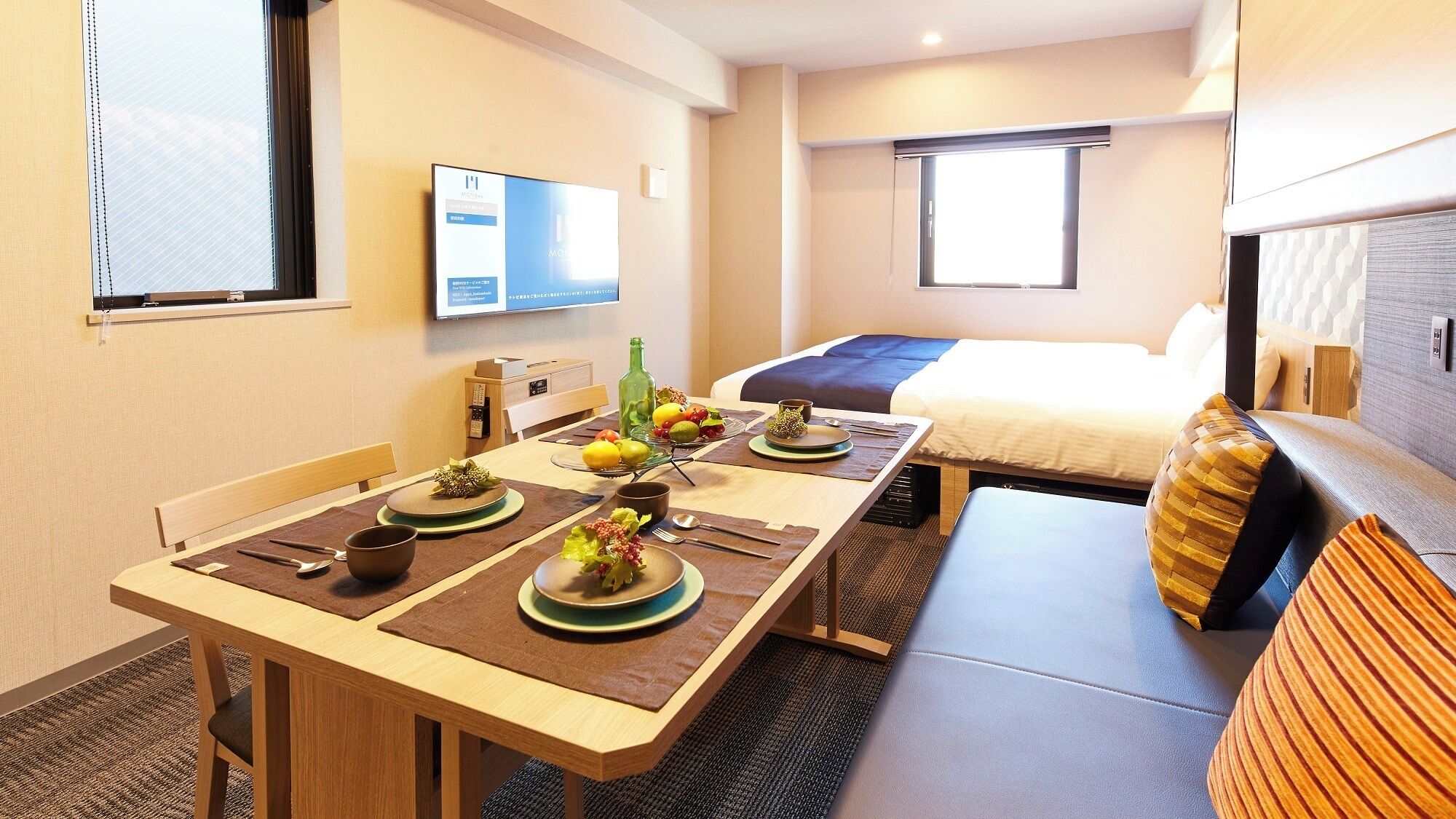 A hotel room with a dining table set for two, a bed, a TV mounted on the wall, and a window with blinds.
