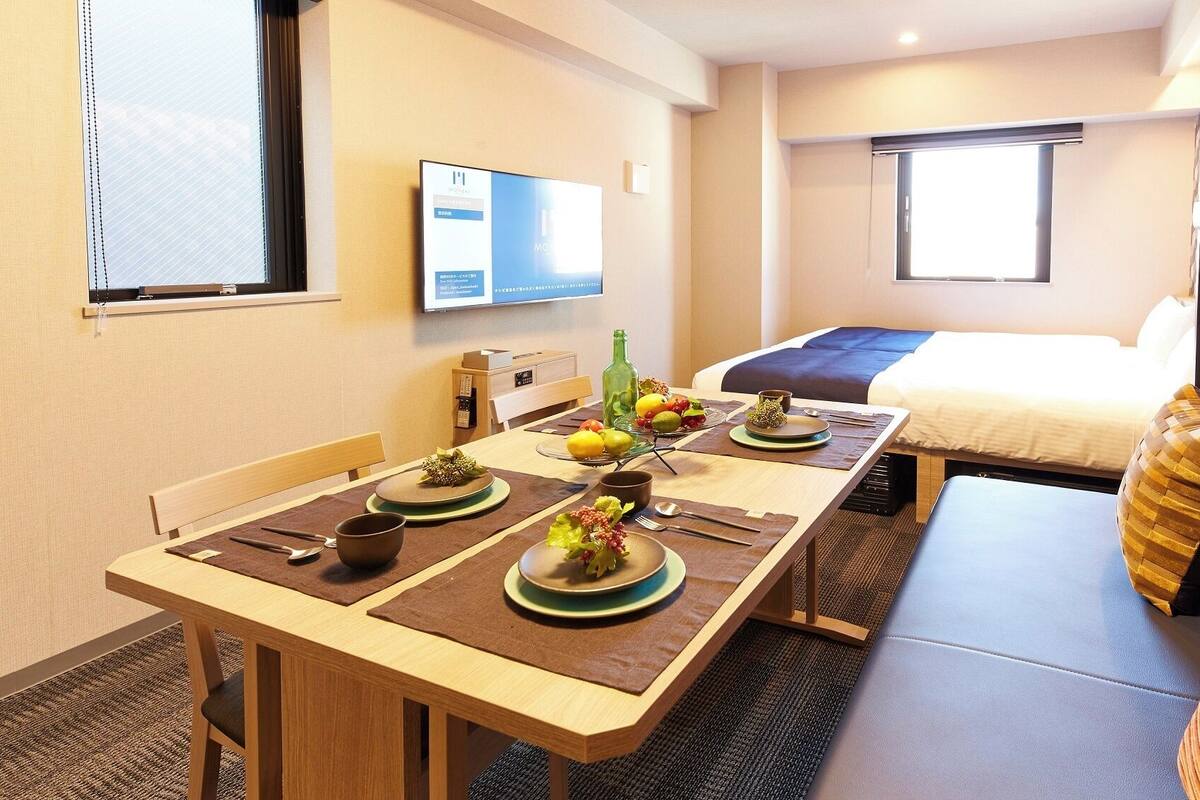 A hotel room with a dining table set for two, a bed, a TV mounted on the wall, and a window with blinds.