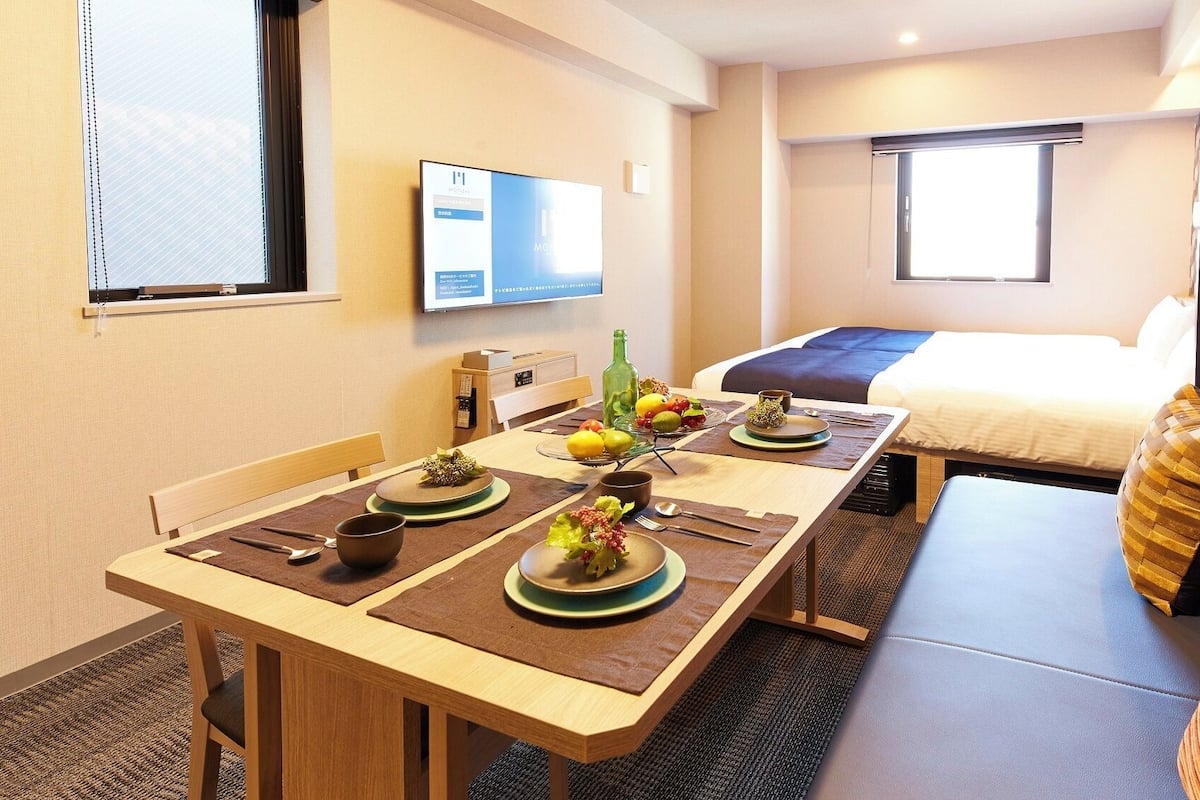 A hotel room with a dining table set for two, a bed, a TV mounted on the wall, and a window with blinds.