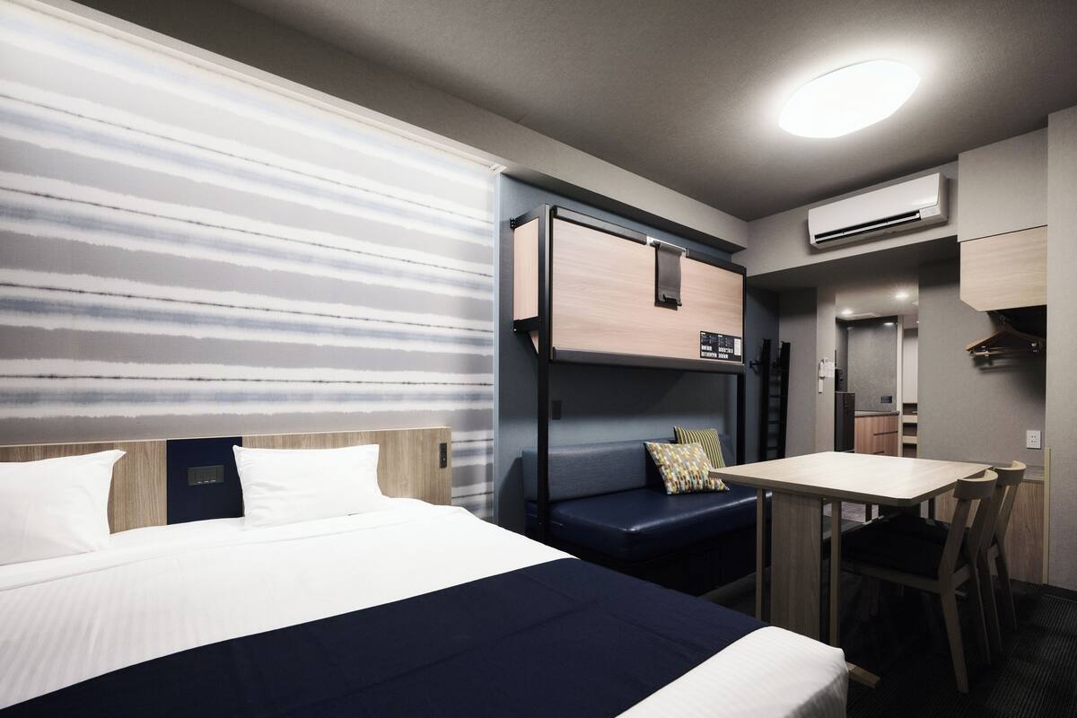 A hotel room with a bed, a desk, a bench, and an air conditioning unit.