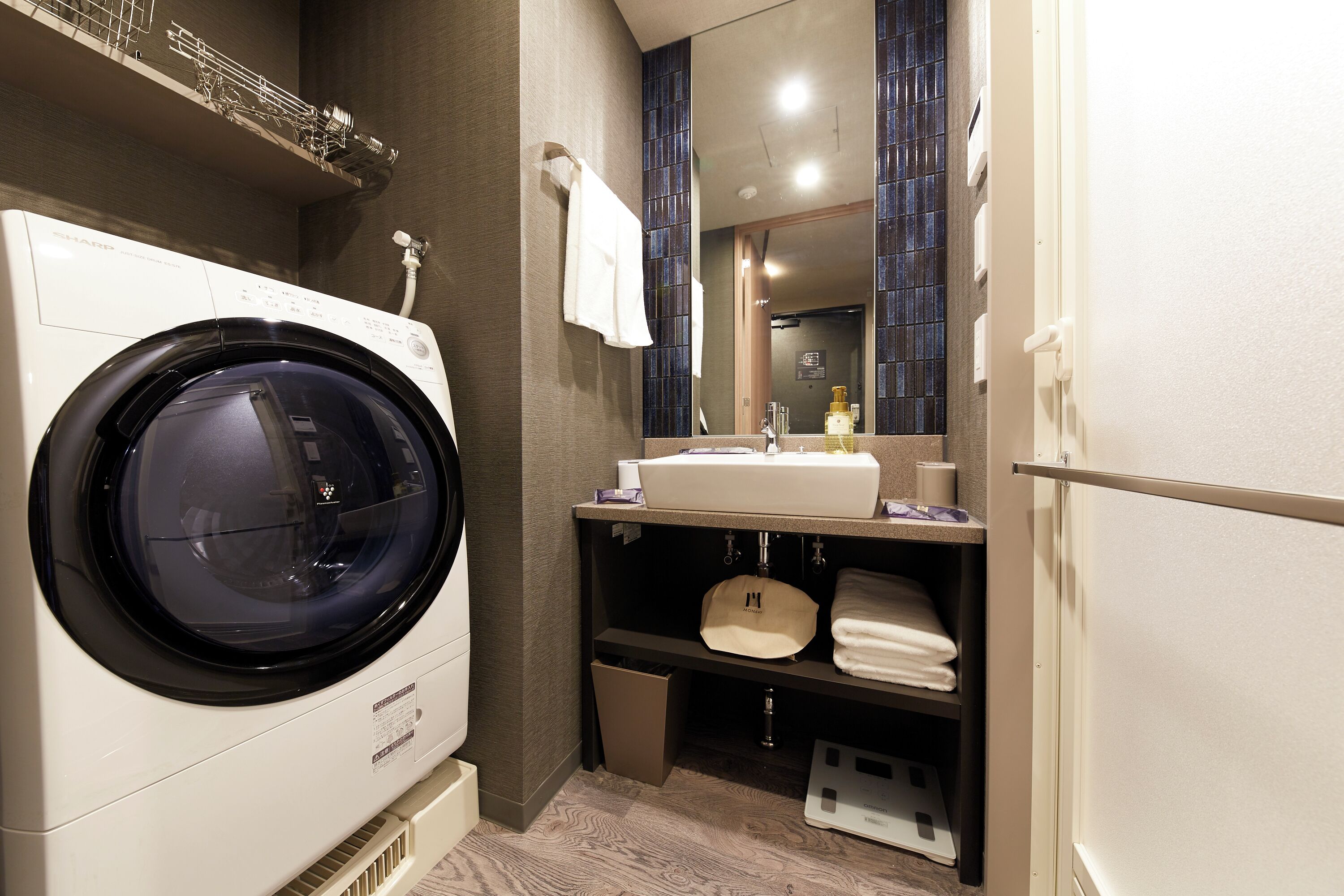A compact hotel bathroom with a front-loading washing machine, a sink with a mirror above, and a towel rack.