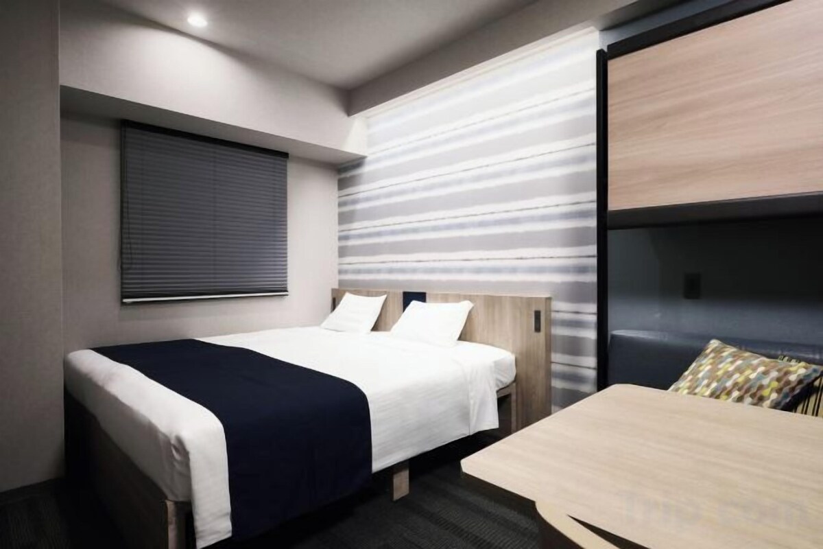 A hotel room with a bed, a small table, a window with blinds, and a wall with a striped pattern.