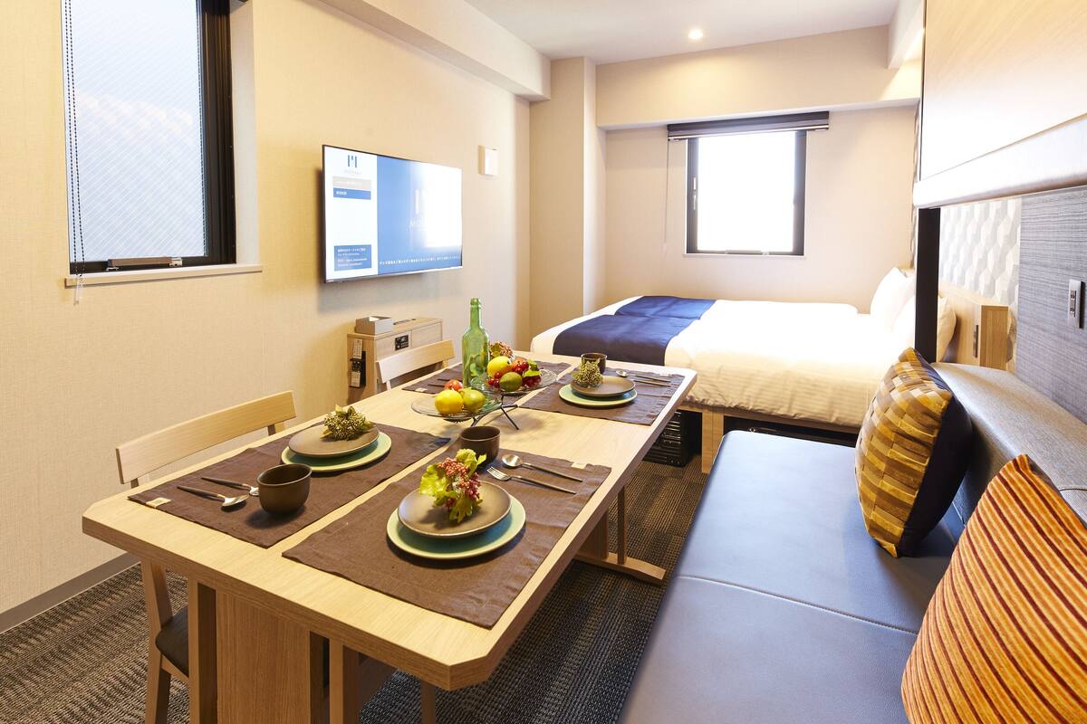 A hotel room with a dining area, a bed, a television, and a window.