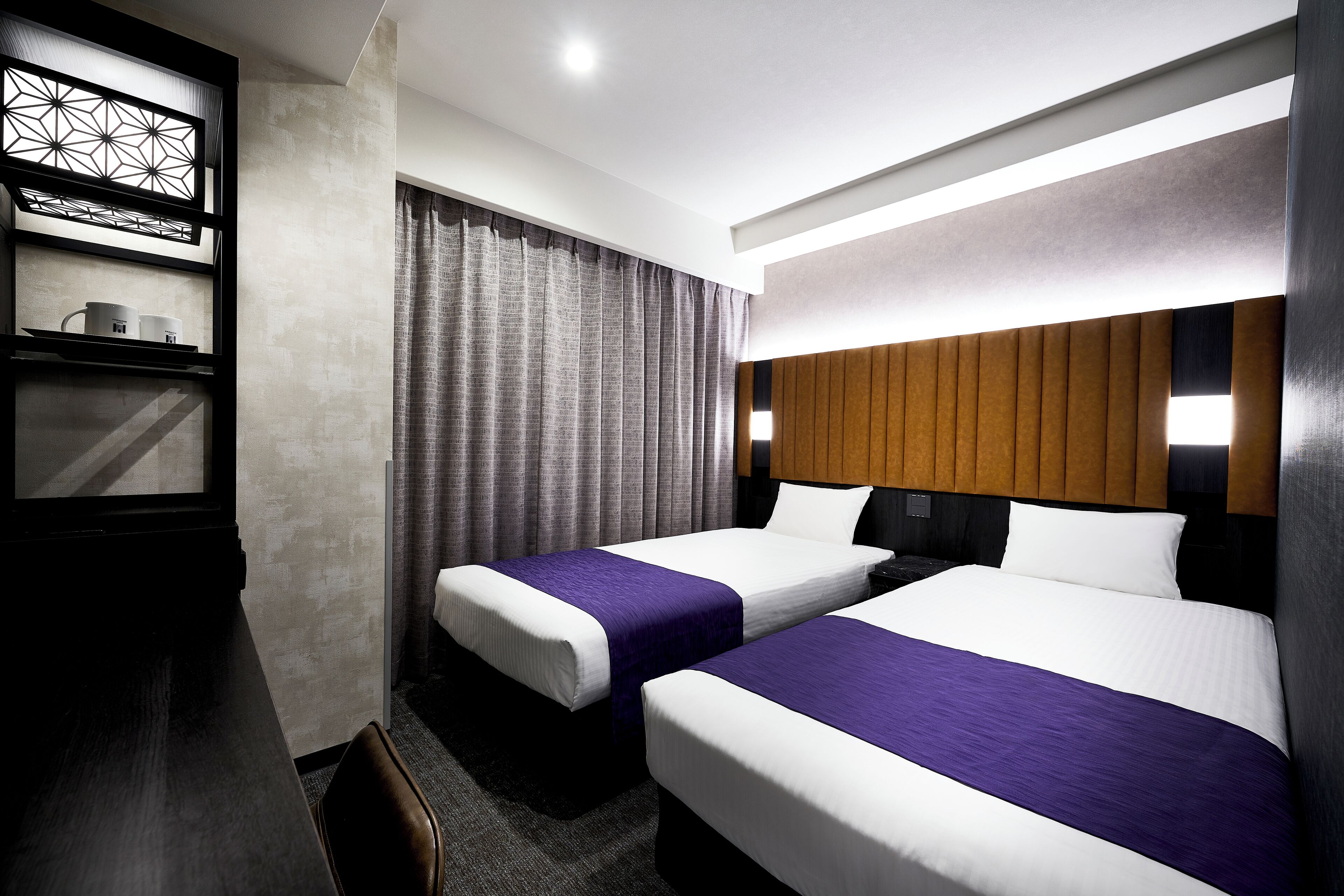 Une chambre d’hôtel avec deux lits, une tête de lit en bois, un bureau avec une chaise, une étagère avec des tasses et une fenêtre avec des rideaux.