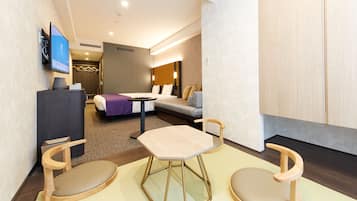 A modern hotel room with a sofa, a small table, two chairs, a TV, and a desk with a lamp.