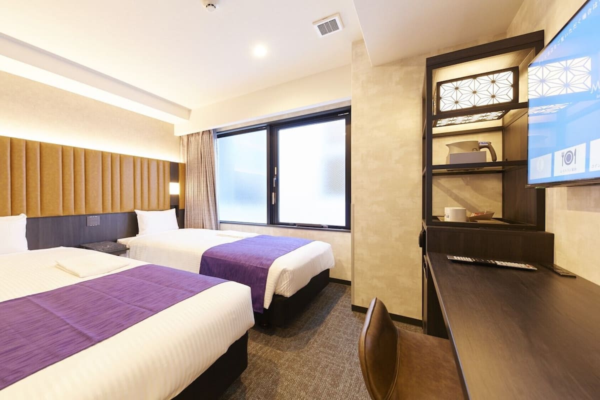 A hotel room with two beds, a desk, a TV, and a window.